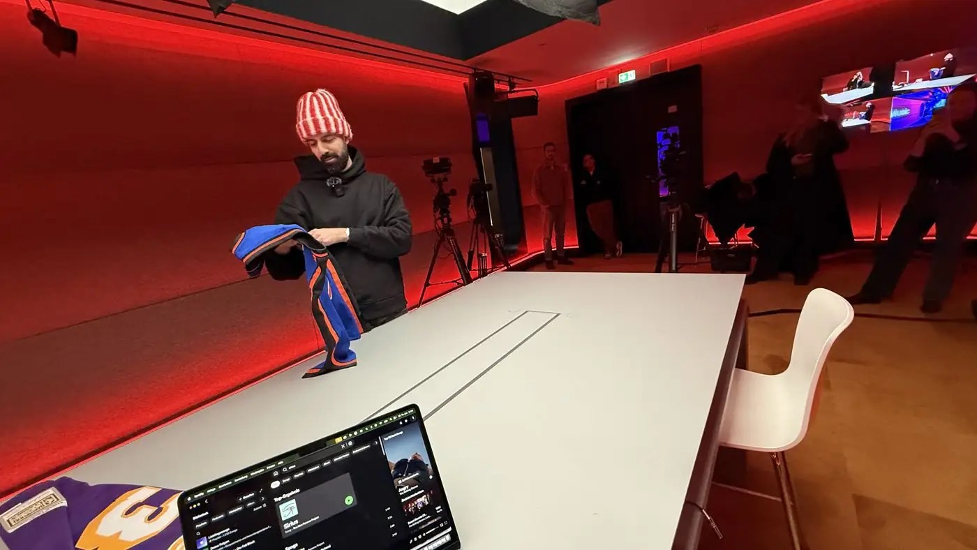 Mann mit gestreifter Mütze faltet ein blau-oranges Trikot in einem rot beleuchteten Studio mit Kameras, vorne ein Tablet mit Musik-App
