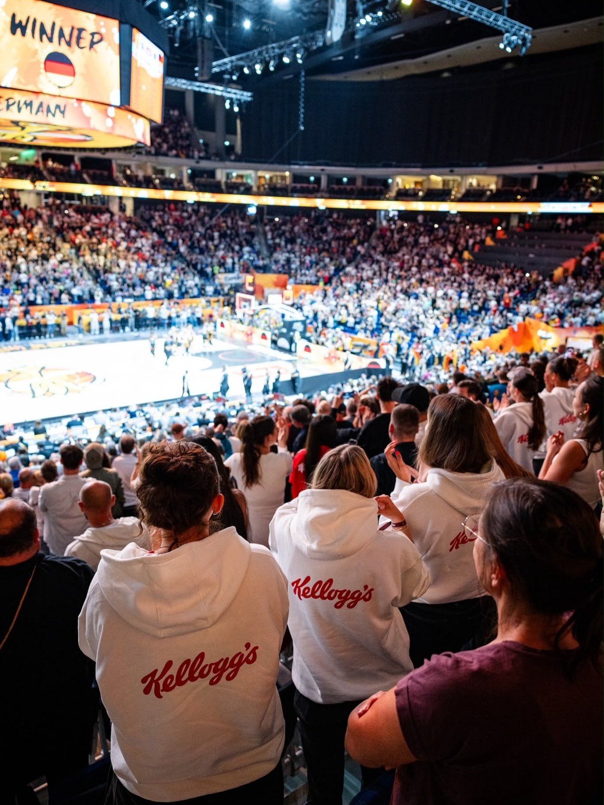 Fans in weißen Kellogg’s-Hoodies stehen auf der Tribüne einer vollen Basketball-Arena und applaudieren, auf dem Videowürfel steht Winner