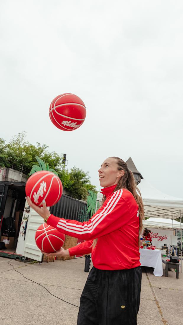 Basketballerin in roter Trainingsjacke jongliert drei rote Kellogg's-Bälle, hinten das Kellogg's-Zelt – Energi