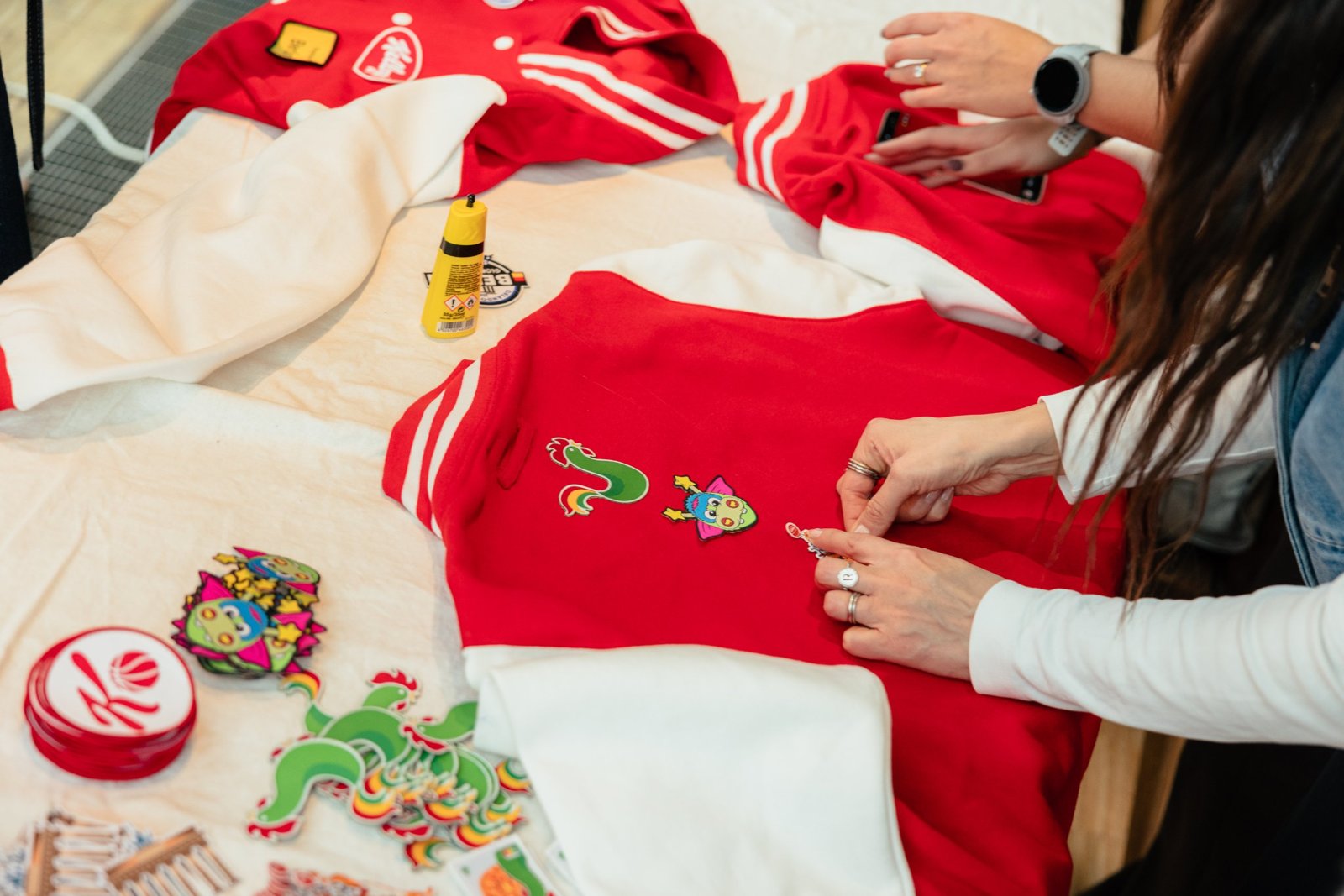 Hände kleben bunte Patches mit Kellogg’s-Figuren auf rote College-Jacken, daneben Stapel weiterer Patches und eine Tube Kleber